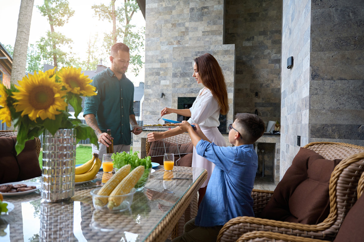 Son helps parents remove hot barbecue food from the grill, sunflowers and fruits on the table